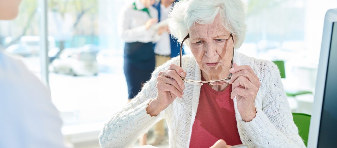 Serious confused senior lady with gray hair sitting at table and looking through eyeglasses while examining document shown by manager in bank office