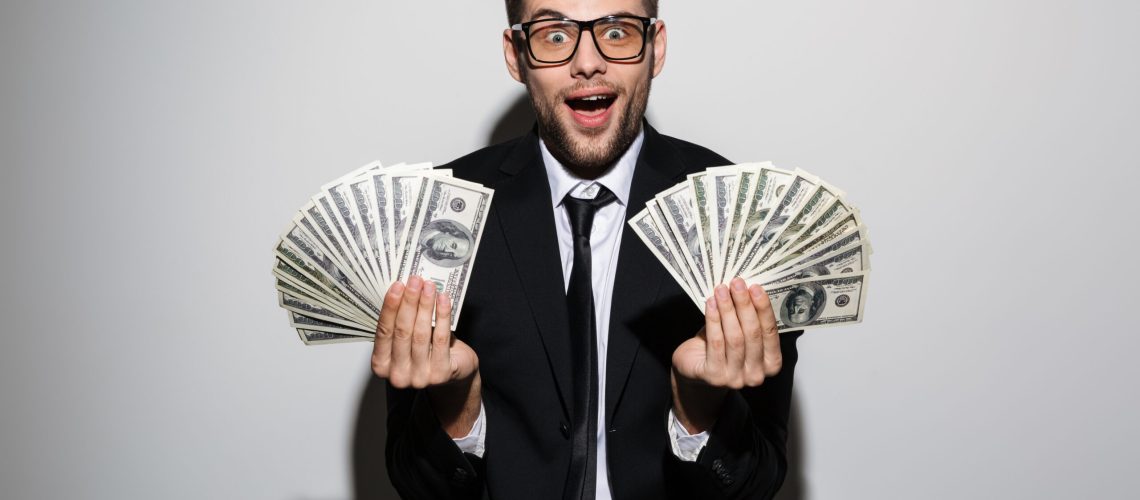 Happy exited man in glasses and black suit holding two bunches of money, looking at camera, isolated over white background