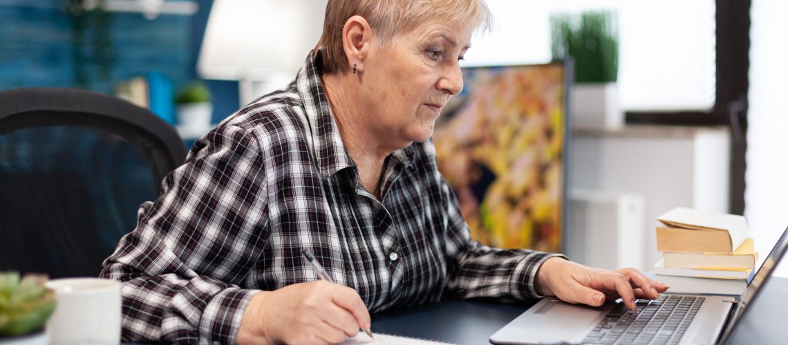 Mature entrepreneur taking notes on notebook working in home office. Elderly woman in home living room using moder technoloy laptop for communication sitting at desk indoors.