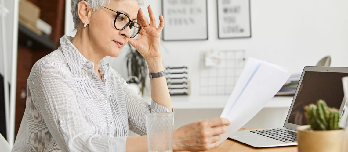 People, aging, technology and profession concept. Serious 50 year old Caucasian female wearing stylish eyeglasses and silk shirt reading contract while working at desk, sitting in front of open laptop